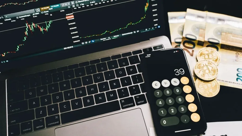 Laptop showing trading charts with calculator, euro bills and Bitcoin coins on a desk